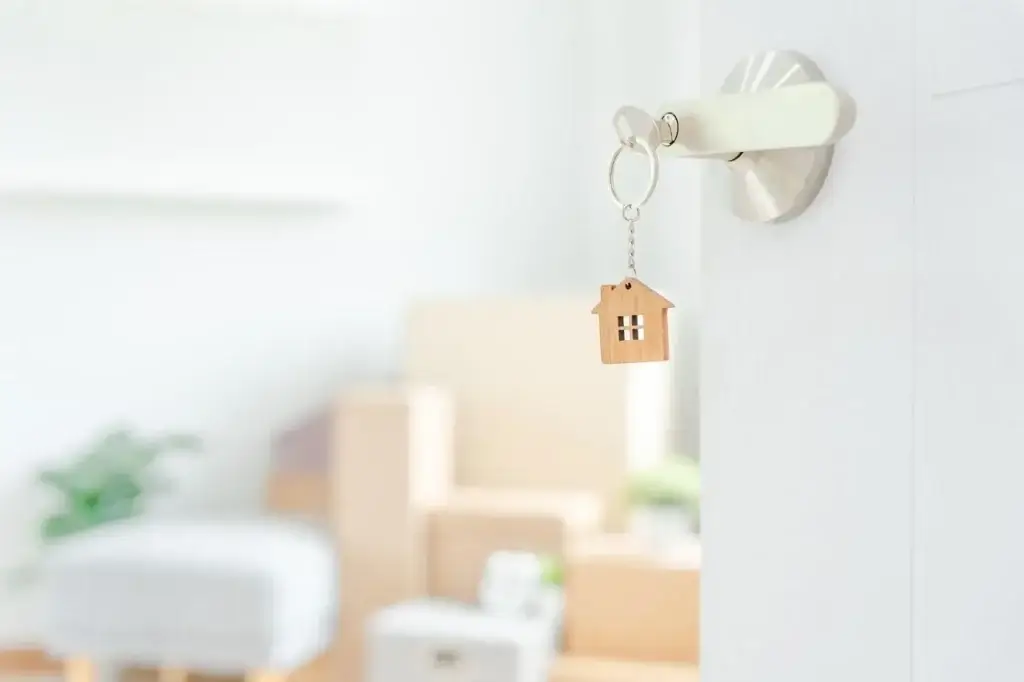 Keys with a wooden house keyring hanging from a door lock, with moving boxes blurred in the background